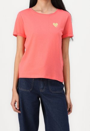 Kvinde iført koralfarvet T-shirt med korte ærmer og lille gyldigt hjerte på brystet samt mørkeblå denim jeans mod en ensfarvet baggrund.