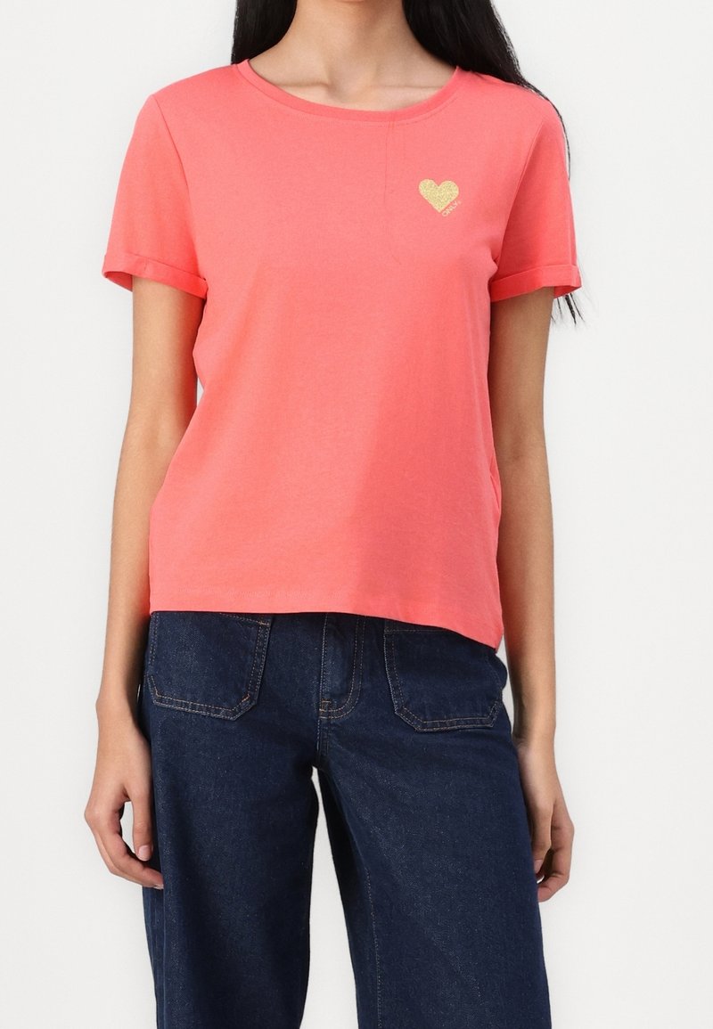 Femme portant un t-shirt corail à manches courtes avec un petit cœur doré sur la poitrine et un jean en denim bleu foncé sur un fond uni.