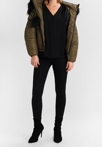 Chaqueta acolchada en verde olivo con detalles de piel sintética negra, combinada con una blusa negra de cuello en V y pantalones ajustados negros, complementada con botas de tacón negras.