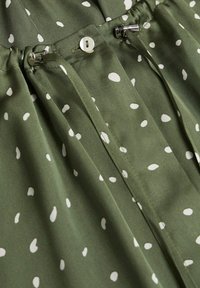 Tessuto verde con pois bianchi, dotato di chiusura con bottone e accessori in metallo. La texture è morbida e presenta dettagli plissettati.