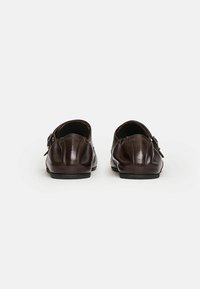 Zapatos de cuero marrón con una textura suave, con una correa trasera y una forma redondeada. Suela plana y un detalle de costura sutil.
