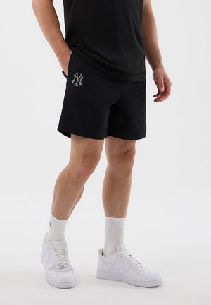 Uomo che indossa pantaloncini neri con un logo bianco sulla coscia sinistra, maglietta nera, calzini bianchi a girocaviglia e scarpe da ginnastica bianche su uno sfondo neutro.