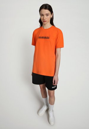 T-shirt di cotone arancione con maniche corte, presenta una stampa nera del logo. Abbinata a pantaloni corti neri e sneakers bianche, mostra un look casual.