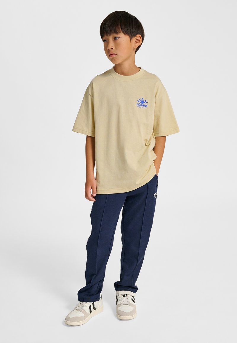 Beżowy bawełniany t-shirt z krótkim rękawem i niebieskim logo, zestawiony z granatowymi spodniami dresowymi. Buty są jasnobeżowe z czarnymi akcentami.