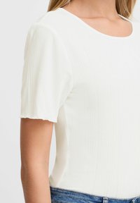 Weißes, geripptes T-Shirt mit kurzen Ärmeln und Rundhalsausschnitt, mit strukturiertem Stoff und rohen Kanten an den Ärmeln. Kombiniert mit blauer Denim.