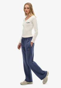 Mujer de pie con una mano en el bolsillo, vestida con una blusa de manga larga crema y pantalones de chándal azules, con zapatillas, sobre fondo blanco.