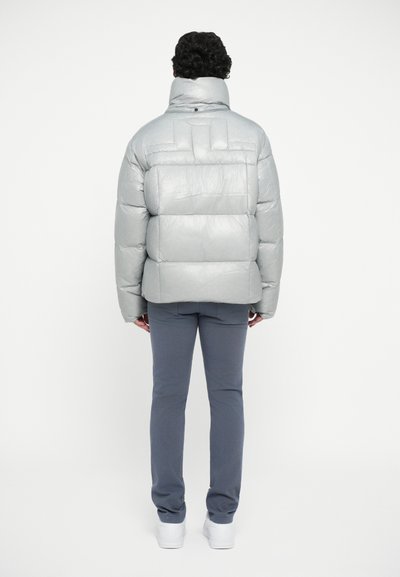 Silver puffer jacket with quilted sections, high collar, and a smooth texture. Paired with dark gray pants and white shoes.
