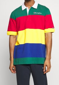 Homme portant un maillot de rugby à manches courtes Champion avec des rayures horizontales vertes, rouges, jaunes, bleues et vertes, avec un col blanc.