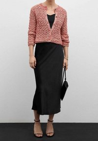 Růžový pletený cardigan přes černé slipové šaty s přiléhavým střihem. Má kulatý výstřih, tříčtvrteční rukávy a strukturovaný vzor.