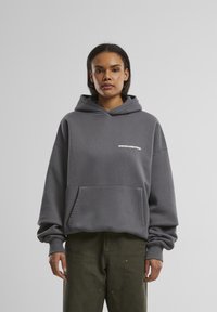 Grijze hoodie met grote voorzak en trekkoordcapuchon, met het woord "PROHIBITED" in het wit op de linker borst. Lange, losse mouwen.