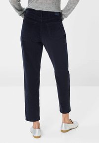 Une personne portant un pantalon en velours côtelé bleu marine, un pull gris et des chaussures plates argentées se tenant contre un fond blanc uni.