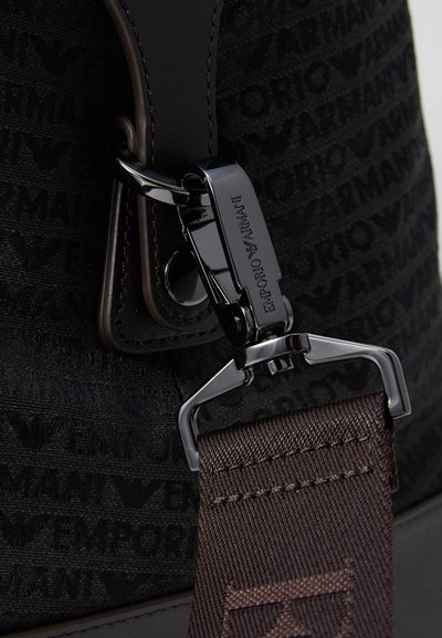 Sac en tissu noir orné d'un motif de logo, rehaussé de cuir marron et d'un fermoir en métal brillant. Anse marron avec une finition texturée.
