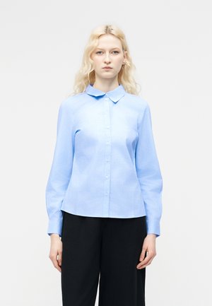 Noor naine pikkade blondide juustega, seljas hele sinine nööpidega pluus ja mustad püksid, seisab ühevärvilise valge tausta ees.