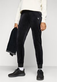 Zwarte fluwelen joggers met ribbels aan de manchetten en een klein logo detail. Model houdt een zwarte handdoek met grijze accenten vast. Draagt zwarte sportschoenen.