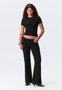 Mlada žena s kovrčavom kosom nosi uski crni crop top, crne široke hlače, crne šiljate cipele i srebrne narukvice, stojeći s povjerenjem.