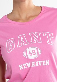 Růžové tričko s bílým nápisem "GANT" zakřiveným nad grafickou ilustrací fotbalu se číslem 49, a "NEW HAVEN" pod ním.