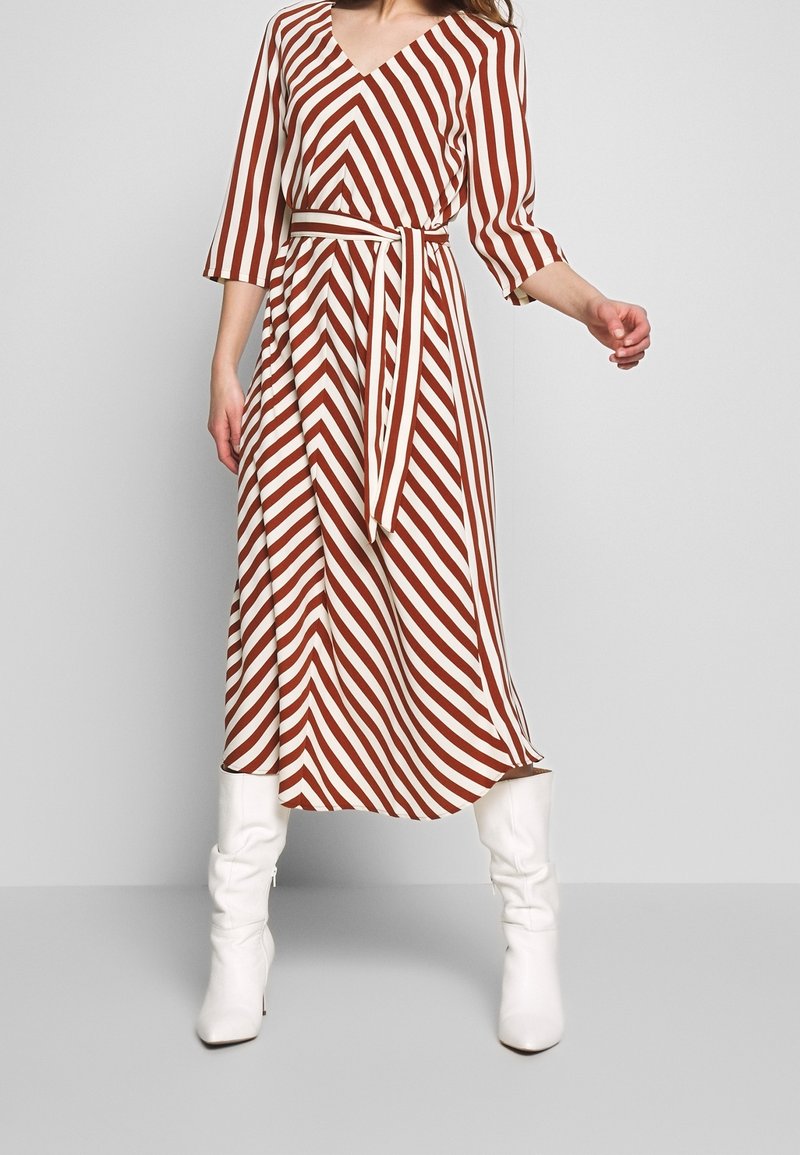 Femme portant une robe midi marron et blanche à rayures diagonales avec une ceinture assortie et des bottes blanches montantes, sur un fond uni.