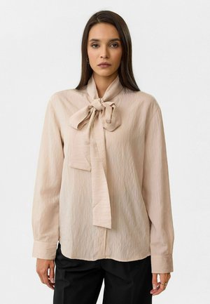 Beige blouse met lange mouwen en een grote strikdetail bij de kraag, gemaakt van een gestructureerde stof. Heeft een relaxte pasvorm en knoopmanchetten.