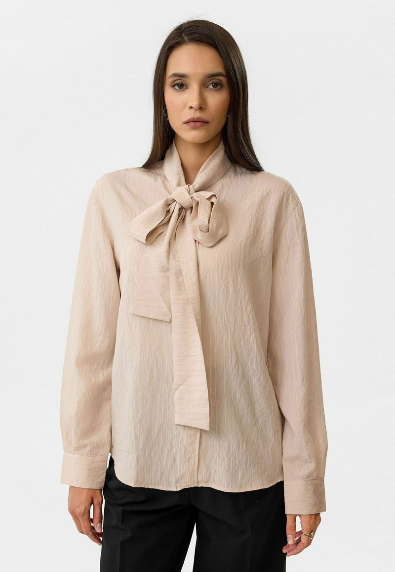 Beige blouse met lange mouwen en een grote strikdetail bij de kraag, gemaakt van een gestructureerde stof. Heeft een relaxte pasvorm en knoopmanchetten.