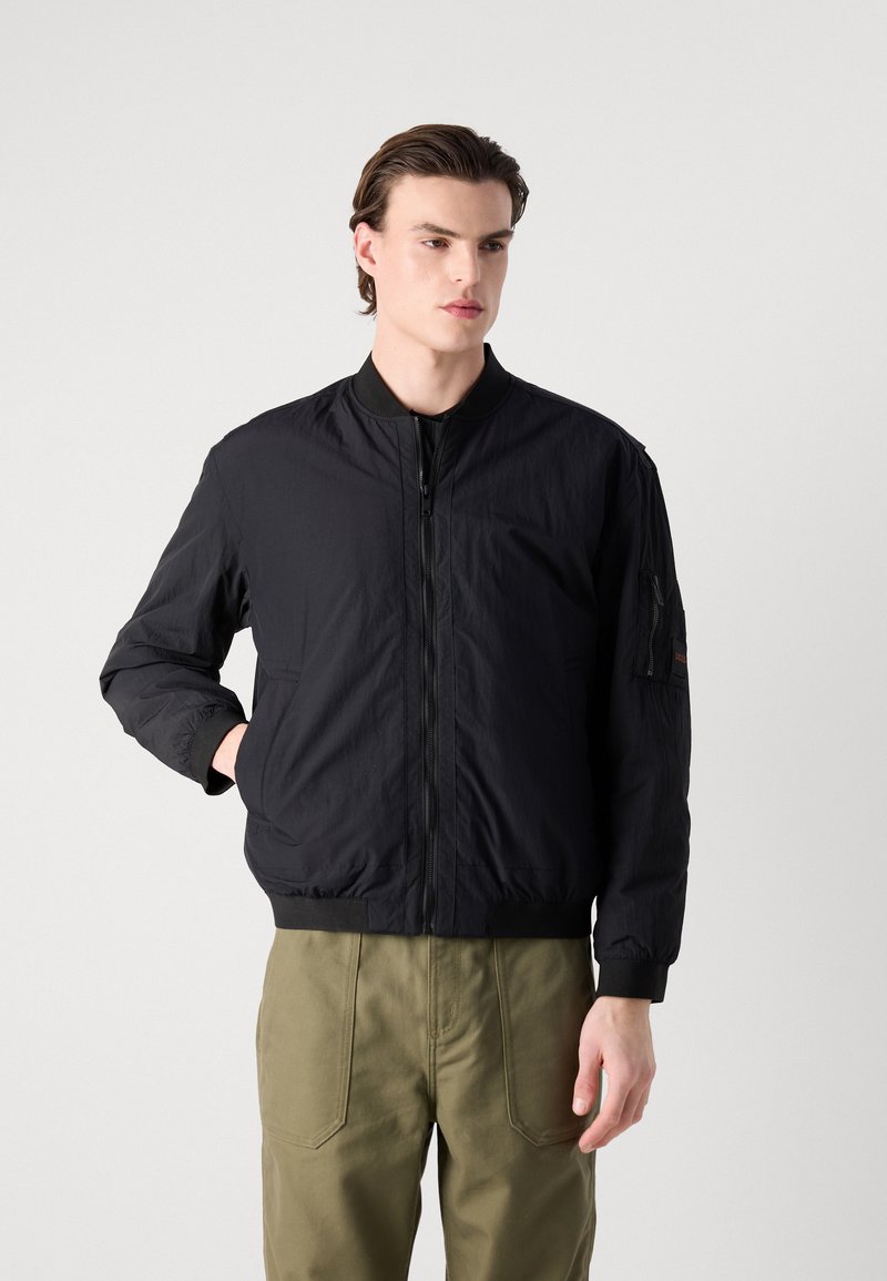 Czarna kurtka bombowiec wykonana z lekkiego materiału, z zapięciem na zamek błyskawiczny z przodu, ściągaczami przy mankietach oraz boczną kieszenią z charakterystycznym przeszyciem.