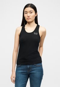 Γυναίκα που φοράει μαύρο αμάνικο τοπ EA7 Emporio Armani και μπλε τζιν, στέκεται μπροστά σε απλό φωτεινό φόντο, κοιτώντας μπροστά.