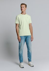 Lichtgroene t-shirt met korte mouwen en ronde hals, gecombineerd met lichtblauwe denim jeans. Het model draagt witte sneakers. Simpel ontwerp.