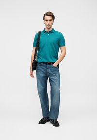 Πετρόλ βαμβακερό πόλο με γιακά, συνδυασμένο με μπλε τζιν παντελόνι και μαύρα παπούτσια. Μια μαύρη τσάντα κρέμεται από τον ώμο, με μίνιμαλ σχεδιασμό.