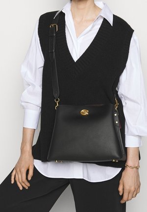 Persoon draagt een zwarte gebreide mouwloze trui over een wit shirt, met een zwarte leren crossbodytas met gouden sluiting en verstelbare riem.