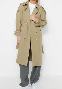 Osoba ubrana w beżowy dwurzędowy trencz na białej koszuli, szarych szerokich spodniach i białych tenisówkach, na tle jednolitego białego tła.