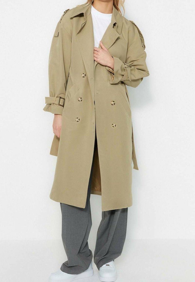 Osoba ubrana w beżowy dwurzędowy trencz na białej koszuli, szarych szerokich spodniach i białych tenisówkach, na tle jednolitego białego tła.