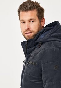 Marineblaue Steppjacke mit Kapuze, ausgestattet mit einer Reißverschlusstasche auf der Brust, strukturiertem Material und schwarzen Hardware-Akzenten.