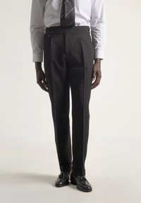 Hombre con pantalones negros a medida, mocasines negros con borlas, camisa blanca a rayas y corbata negra a rayas, contra un fondo liso.