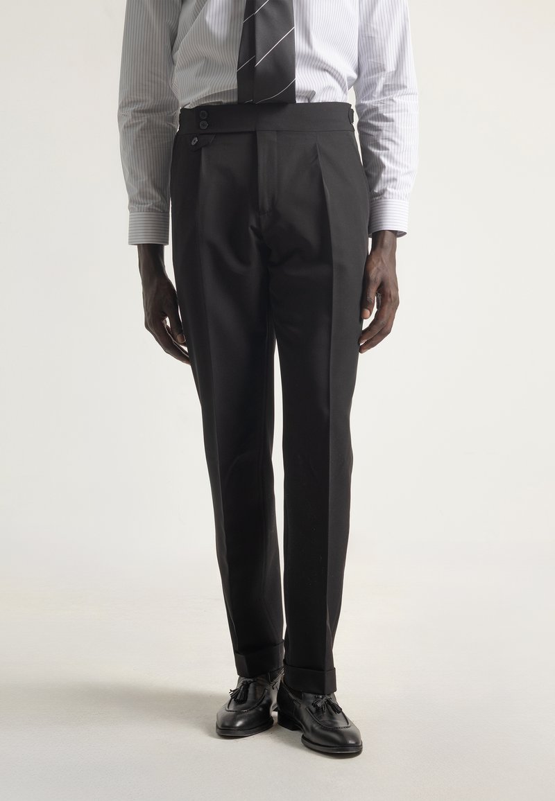 Hombre con pantalones negros a medida, mocasines negros con borlas, camisa blanca a rayas y corbata negra a rayas, contra un fondo liso.