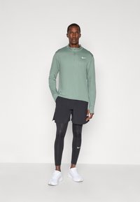 Langärmliges grünes Sportoberteil, schwarze Shorts und schwarze Leggings, mit weißen Nike-Logo-Akzenten und weißen Sportschuhen.