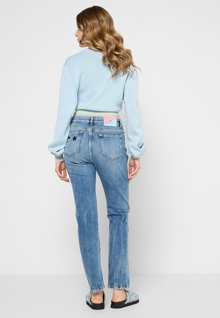 Vrouw met golvend haar, gekleed in een lichtblauwe trui, hoge blauwe jeans met patches en lichtblauwe sandalen, staande met haar rug naar de camera op een witte achtergrond.