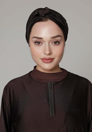 Femme à la peau claire porte un turban noir torsadé et un haut zippé sombre, face à l'avant avec une expression neutre sur un fond uni.