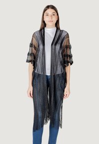 Kimono nero a maglia con maniche ampie, caratterizzato da un orlo a frange e design aperto sul davanti. Indossato sopra una maglietta bianca e jeans blu.