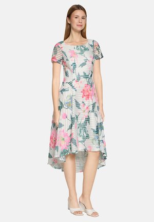 Frau trägt ein weißes High-Low-Kleid mit rosa und grünen Blumen, kurzen Ärmeln und weißen offenen Blockabsatz-Sandalen.