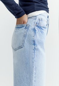 Persoană purtând blugi albastru deschis și pulover bleumarin, cu o mână pusă în buzunarul din spate, pe un fundal alb simplu.