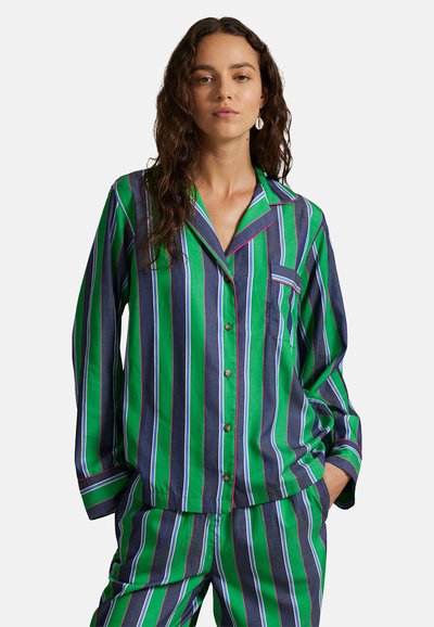 Frau trägt einen grün-navyblau vertikal gestreiften Pyjama mit Knöpfen und langen Ärmeln, steht mit den Händen in den Taschen.