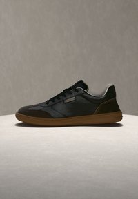 Basket noire basse avec semelle marron, devant à lacets et doublure intérieure blanche sur une surface claire et un fond neutre.