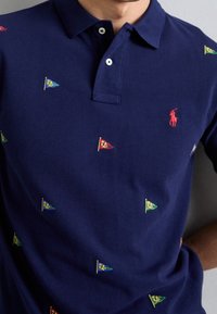 Námořnická modrá polokošile z texturované bavlny, zdobená barevnými vyšitými vlajkami a malým červeným logem na hrudi.