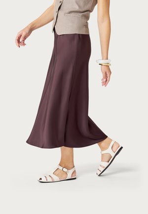 Mujer caminando con un chaleco sin mangas beige, falda midi morado oscuro, sandalias blancas planas y pulseras gruesas blancas y doradas.