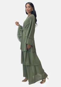 Olivgrünes, mehrlagiges Maxikleid mit weiten Ärmeln und Bindegürtel. Transparenter, fließender Stoff und bodenlanger Saum. Silberne Sandalen mit Absatz.