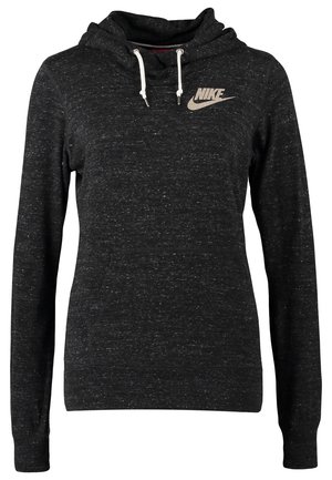 Osoba stojąca w czarnej bluzie z kapturem Nike, niebieskich obcisłych dżinsach, granatowych tenisówkach i nosząca czarny plecak na jednym ramieniu.