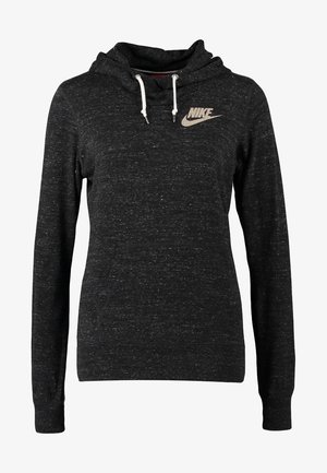 Osoba stojąca w czarnej bluzie z kapturem Nike, niebieskich obcisłych dżinsach, granatowych tenisówkach i nosząca czarny plecak na jednym ramieniu.