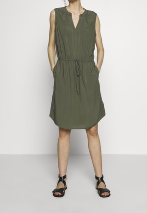 Femme debout, les mains dans les poches, portant une robe sans manches vert olive longueur genou et des sandales noires à brides croisées sur un fond uni.