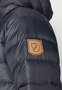 Mornarska puf jakna s kvadriranim dijelovima, izrađena od sjajnog materijala. Uključuje bež kožni oznaku s logotipom lisice na rukavu.