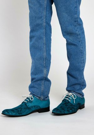 Teal gekleurde dresschoenen met een gladde textuur, zwarte zool en witte veters, gedragen met een spijkerbroek.