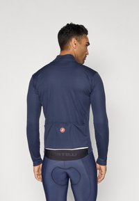 Mann, der ein langärmeliges Fahrradtrikot in Marineblau und gepolsterte Fahrradhosen trägt, von hinten zu sehen, vor einem schlichten hellen Hintergrund.